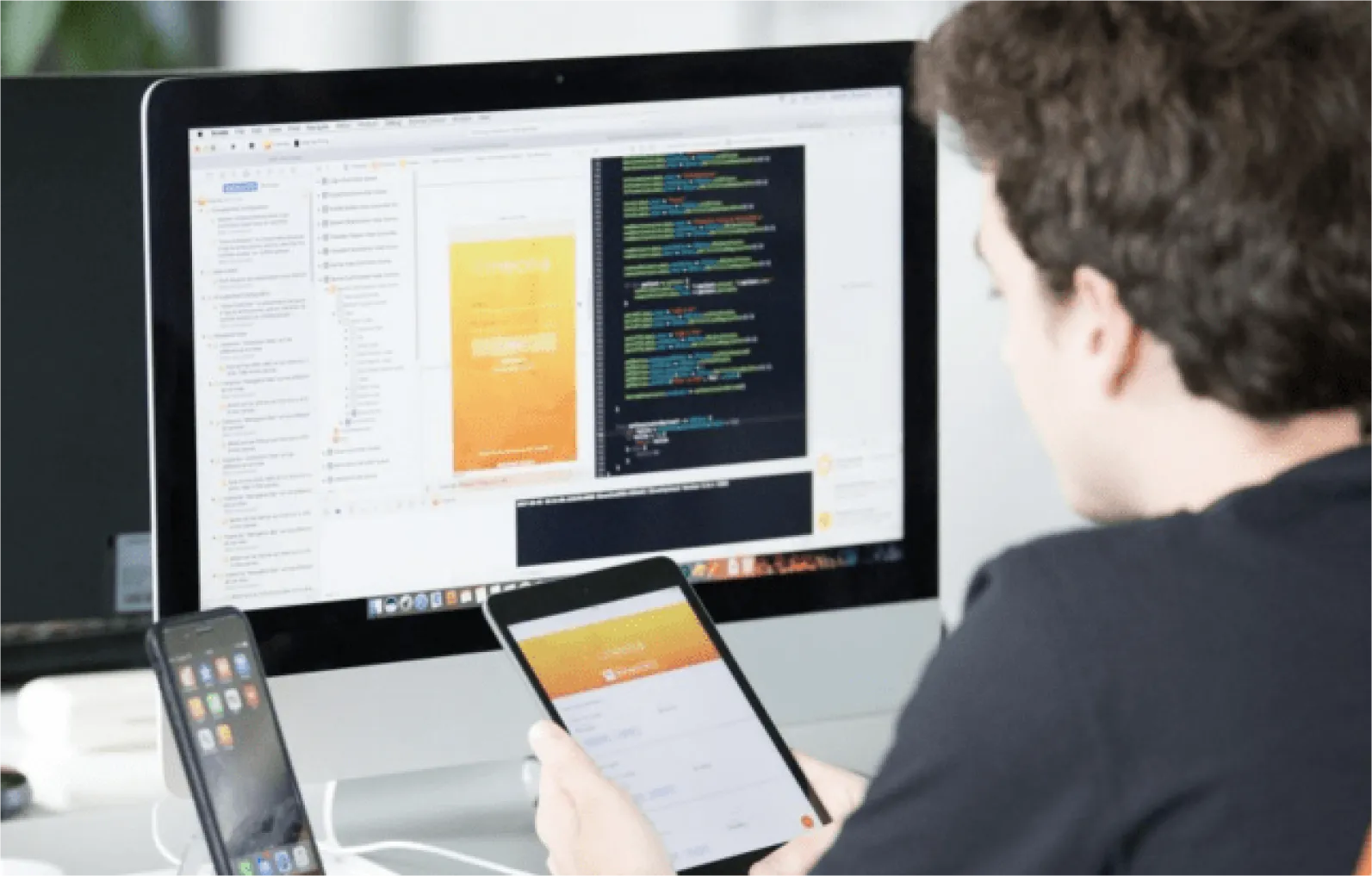 Person holding a tablet displaying an app interface while looking at a computer screen showing code and app design.