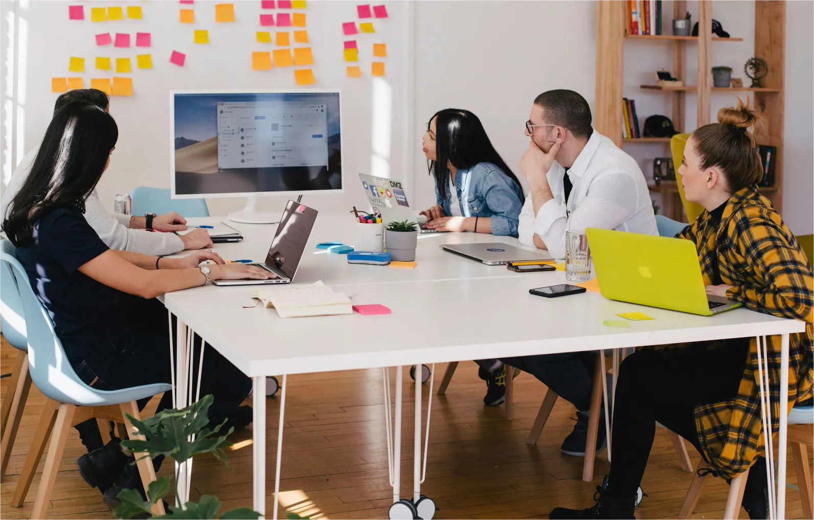 Five people sitting around a white table in a meeting room, looking at a computer monitor with sticky notes on the wall behind them.